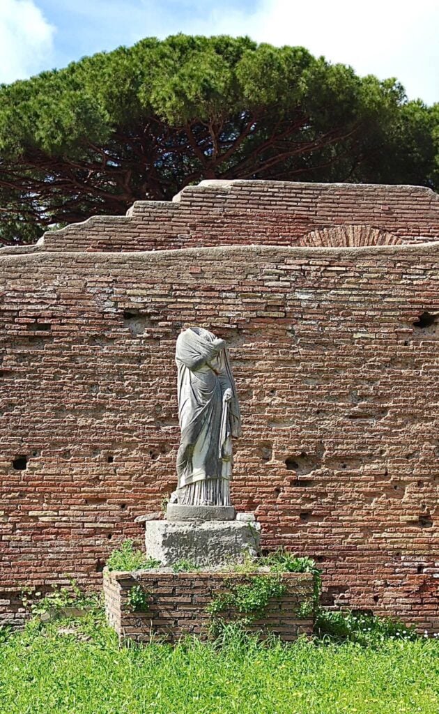 A cast of a Roman statue standing outdoors against the brick archaeological remains of Ostia Antica.